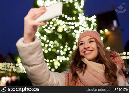 holidays and people concept - beautiful happy young woman taking selfie over christmas tree lights in winter evening. young woman taking selfie over christmas tree