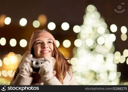 holidays and people concept - beautiful happy young woman with camera over christmas tree lights in winter evening. happy young woman with camera over christmas tree