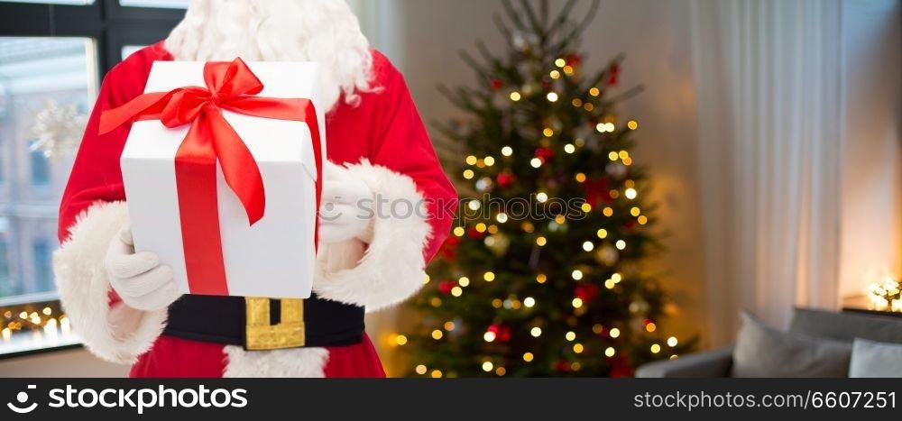holidays and people concept - close up of santa claus with gift box over room with christmas tree background. santa claus with christmas gift over room