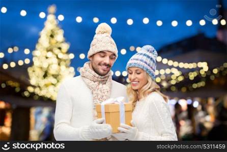 holidays and people concept - happy couple in winter hats with gift box over christmas tree lights background. happy couple with gift box over christmas lights