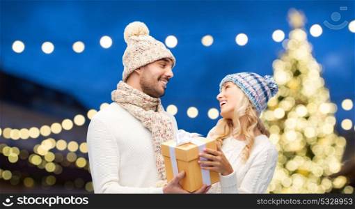 holidays and people concept - happy couple in winter hats with gift box over christmas tree lights background. happy couple with gift box over christmas lights