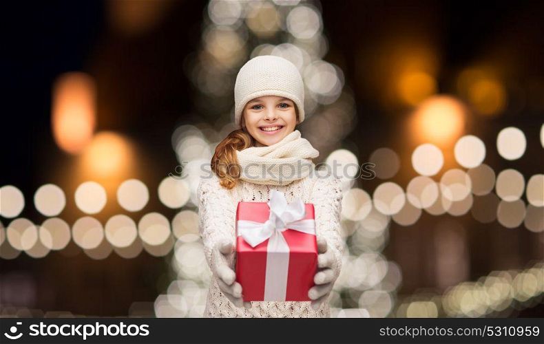 holidays and people concept - happy girl in winter clothes with gift box over christmas tree lights background. happy girl in winter clothes with gift box