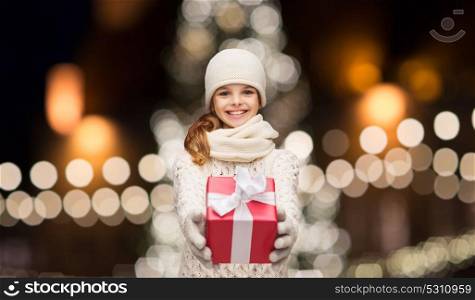 holidays and people concept - happy girl in winter clothes with gift box over christmas tree lights background. happy girl in winter clothes with gift box
