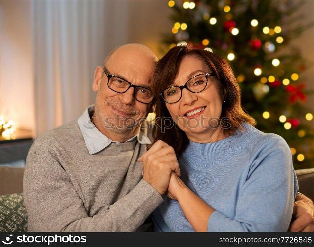 holidays and people concept - happy smiling senior couple hugging on sofa at home in evening over christmas tree lights on background. happy senior couple hugging at home on christmas