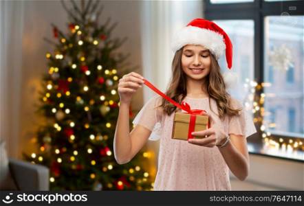 holidays and people concept - happy smiling teenage girl in santa helper hat opening gift box over christmas tree lights background. teenage girl in santa hat opening christmas gift