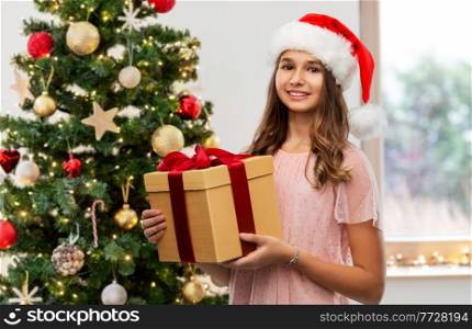 holidays and people concept - happy smiling teenage girl in santa helper hat holding gift box over christmas tree on background. teenage girl in santa hat with christmas gift