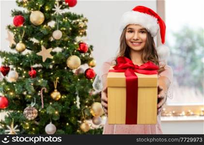 holidays and people concept - happy smiling teenage girl in santa helper hat holding gift box over christmas tree on background. teenage girl in santa hat with christmas gift