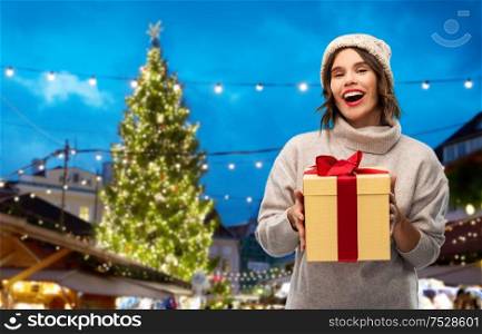 holidays and people concept - happy smiling young woman in knitted winter hat and sweater holding gift box over christmas tree at old town square market in tallinn, estonia background. woman in hat holding gift box at christmas market