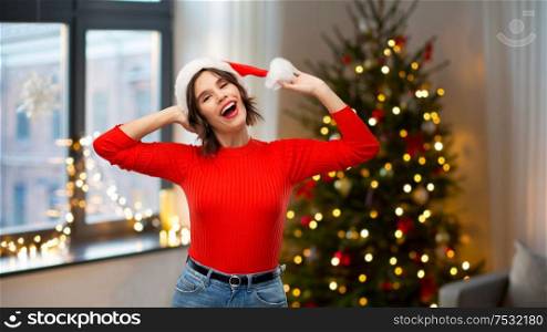 holidays and people concept - happy smiling young woman in santa helper hat over christmas tree lights at home background. happy young woman in santa hat on christmas