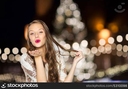 holidays and people concept - happy young woman or teen girl in fancy dress sending air kiss over christmas tree lights background. happy woman sending air kiss over christmas lights