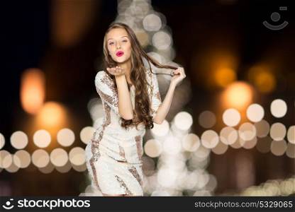 holidays and people concept - happy young woman or teen girl in fancy dress sending air kiss over christmas tree lights background. happy woman sending air kiss over christmas lights