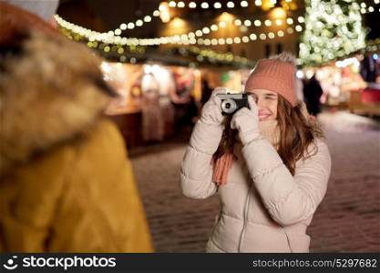holidays and people concept - happy young woman with camera photographing her boyfriend at christmas market in winter evening. woman with camera photographing man at christmas