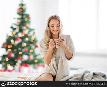 holidays and people concept - happy young woman with cup of coffee or tea in bed at home bedroom over christmas tree background. happy woman with cup of coffee in bed at christmas