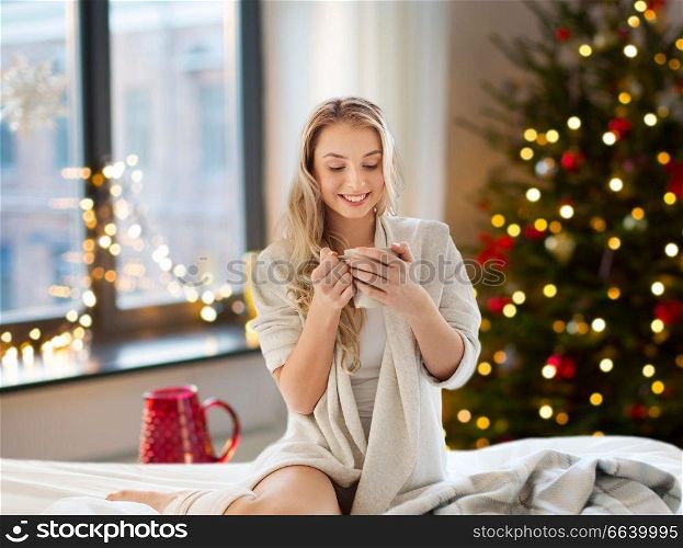 holidays and people concept - happy young woman with cup of coffee or tea in bed at home bedroom over christmas tree lights on background. woman drinking coffee in bed at home on christmas