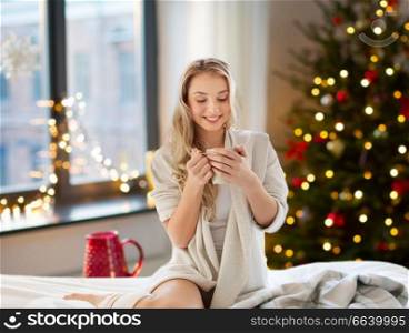 holidays and people concept - happy young woman with cup of coffee or tea in bed at home bedroom over christmas tree lights on background. woman drinking coffee in bed at home on christmas