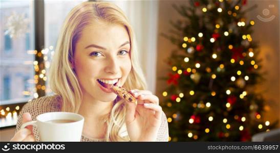 holidays and people concept - happy young woman with cup of tea eating cookie at home over christmas tree lights on background. woman with tea eating cookie on christmas at home