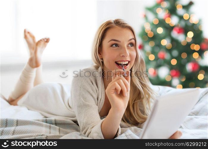 holidays and people concept - happy young woman with pen and notebook lying in bed and writing at home over christmas tree background. happy woman with notebook in bed at christmas