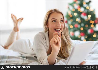 holidays and people concept - happy young woman with pen and notebook lying in bed and writing at home over christmas tree background. happy woman with notebook in bed at christmas
