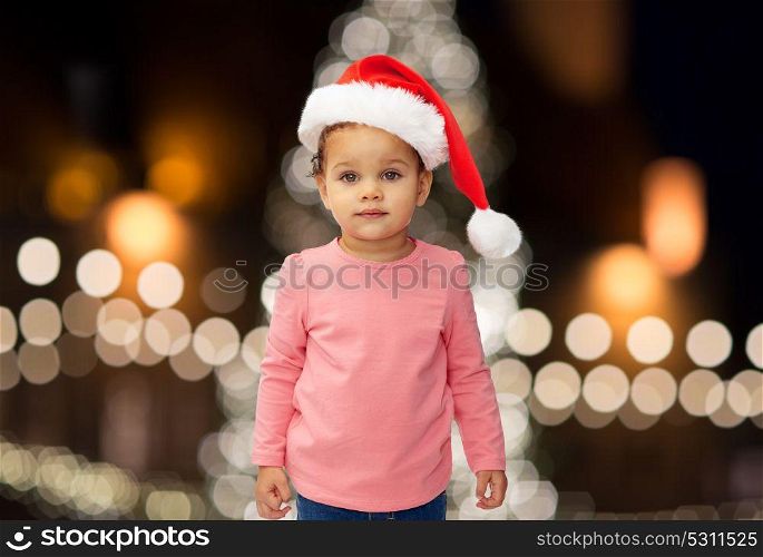 holidays and people concept - little baby girl in santa hat over christmas tree lights background. little baby girl in santa hat at christmas
