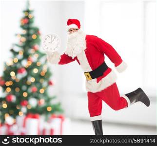 holidays and people concept - man in costume of santa claus running with clock showing twelve over living room and christmas tree background