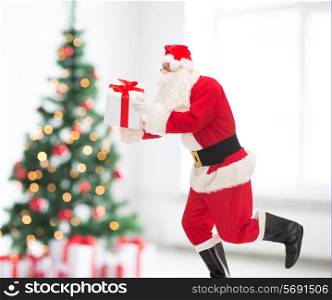 holidays and people concept - man in costume of santa claus running with gift box over living room and christmas tree background