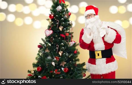 holidays and people concept - man in costume of santa claus with bag and christmas tree making hush gesture. santa claus with bag and christmas tree
