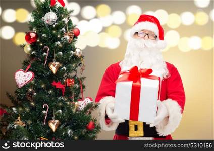 holidays and people concept - man in costume of santa claus with gift box at christmas tree over lights background. santa claus with gift box at christmas tree