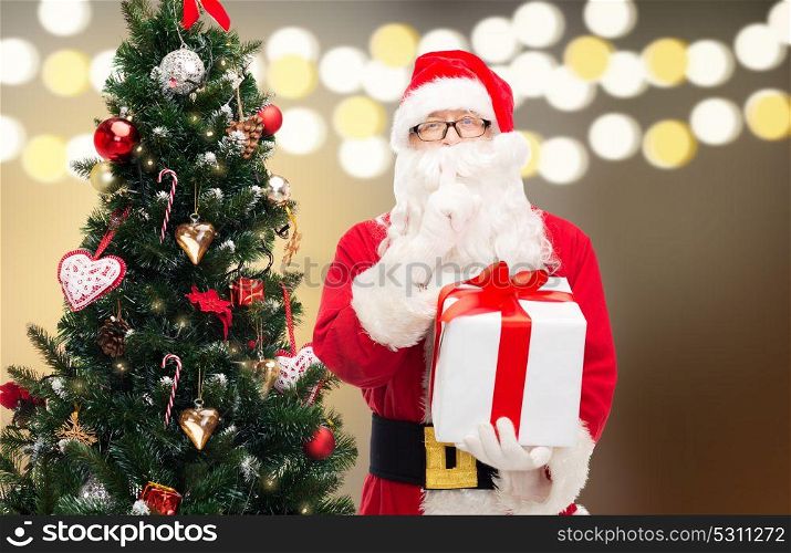 holidays and people concept - man in costume of santa claus with gift box and christmas tree over lights background making hush gesture. santa claus with gift box at christmas tree