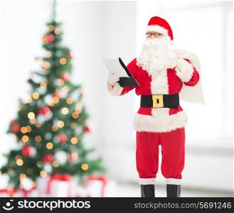 holidays and people concept - man in costume of santa claus with notepad and bag over living room and christmas tree background