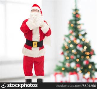 holidays and people concept - man in costume of santa claus with bag making hush gesture over living room and christmas tree background