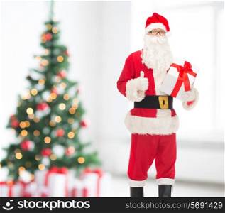holidays and people concept - man in costume of santa claus with gift box showing thumbs up gesture over living room and christmas tree background