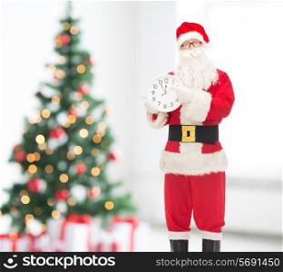 holidays and people concept - man in costume of santa claus with clock showing twelve pointing finger over living room and christmas tree background