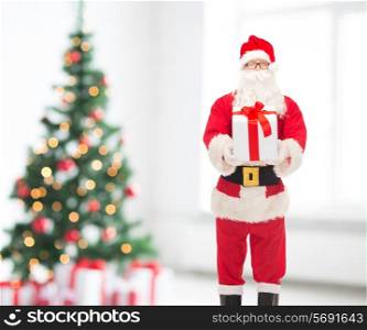 holidays and people concept - man in costume of santa claus with gift box over living room and christmas tree background