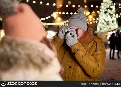 holidays and people concept - man with camera photographing his girlfriend at christmas market in winter evening. man with camera photographing woman at christmas