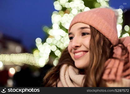 holidays and people concept - portrait of beautiful happy young woman over christmas tree lights in winter evening. happy young woman at christmas market in winter