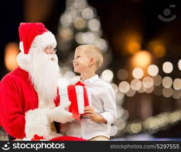 holidays and people concept - santa claus and happy little boy with gift box over christmas tree lights background. santa claus and happy boy with christmas gift
