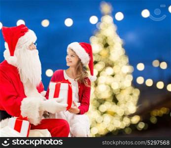 holidays and people concept - santa claus and happy little girl with gift box over christmas tree lights background. santa claus and happy girl with christmas gift