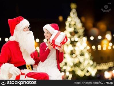holidays and people concept - santa claus and happy little girl with gift box over christmas tree lights background. santa claus and happy girl with christmas gift
