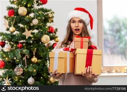 holidays and people concept - shocked teenage girl in santa helper hat holding gift box over christmas tree on background. teenage girl in santa hat with christmas gift