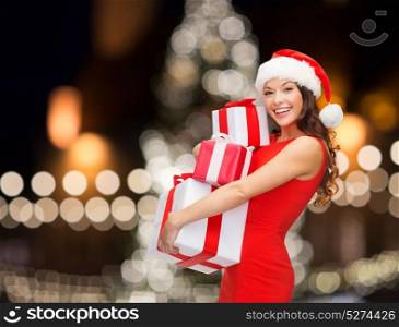 holidays and people concept- smiling woman in santa hat with gift boxes over night christmas tree lights background. smiling woman in santa hat with gift boxes