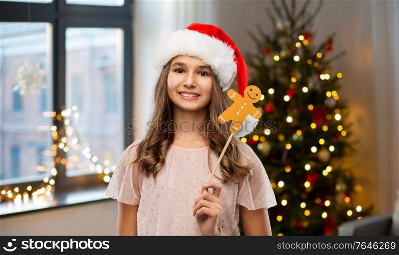 holidays and photo booth concept - happy smiling teenage girl in santa helper hat with gingerbread man party accessory over christmas tree lights background. happy teenage girl in santa hat on christmas