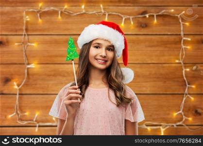 holidays and photo booth concept - happy smiling teenage girl in santa helper hat with christmas tree party accessory over garland lights on wooden background. happy teenage girl in santa hat on christmas