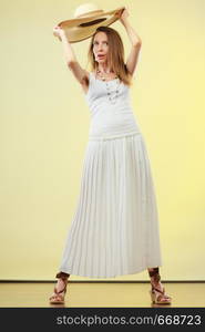 Holidays and summer fashion. Woman wearing big straw hat white dress. Female model posing in full length on bright yellow background.