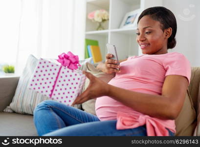 holidays and technology concept - happy pregnant african american woman sitting on sofa at home and taking picture of gift box with smartphone. african pregnant woman with smartphone and gift