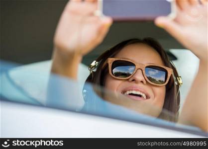 Holidays and tourism concept - smiling teenage girl taking selfie picture with smartphone camera outdoors in car