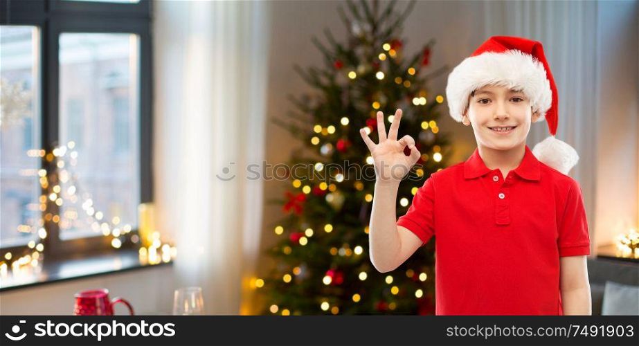 holidays and winter childhood concept - smiling little boy in santa helper hat showing ok hand sign over christmas tree at home background. smiling boy in santa helper hat showing ok gesture