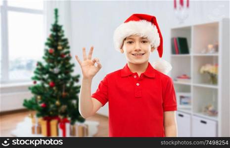 holidays and winter childhood concept - smiling little boy in santa helper hat showing ok hand sign over christmas tree at home background. smiling boy in santa helper hat showing ok gesture