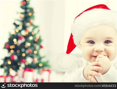 holidays, babyhood, childhood and people concept - happy baby in santa hat over christmas tree lights and gifts background