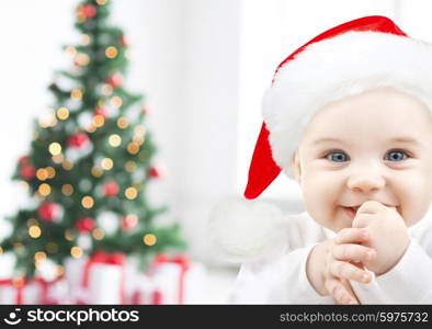 holidays, babyhood, childhood and people concept - happy baby in santa hat over christmas tree lights and gifts background