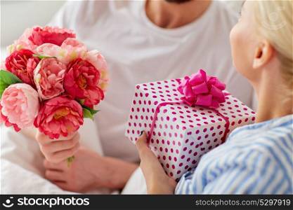 holidays, birthday, valentines day, romance and people concept - close up of happy couple with gift box and flowers in bed at home. close up of couple with gift and flowers in bed